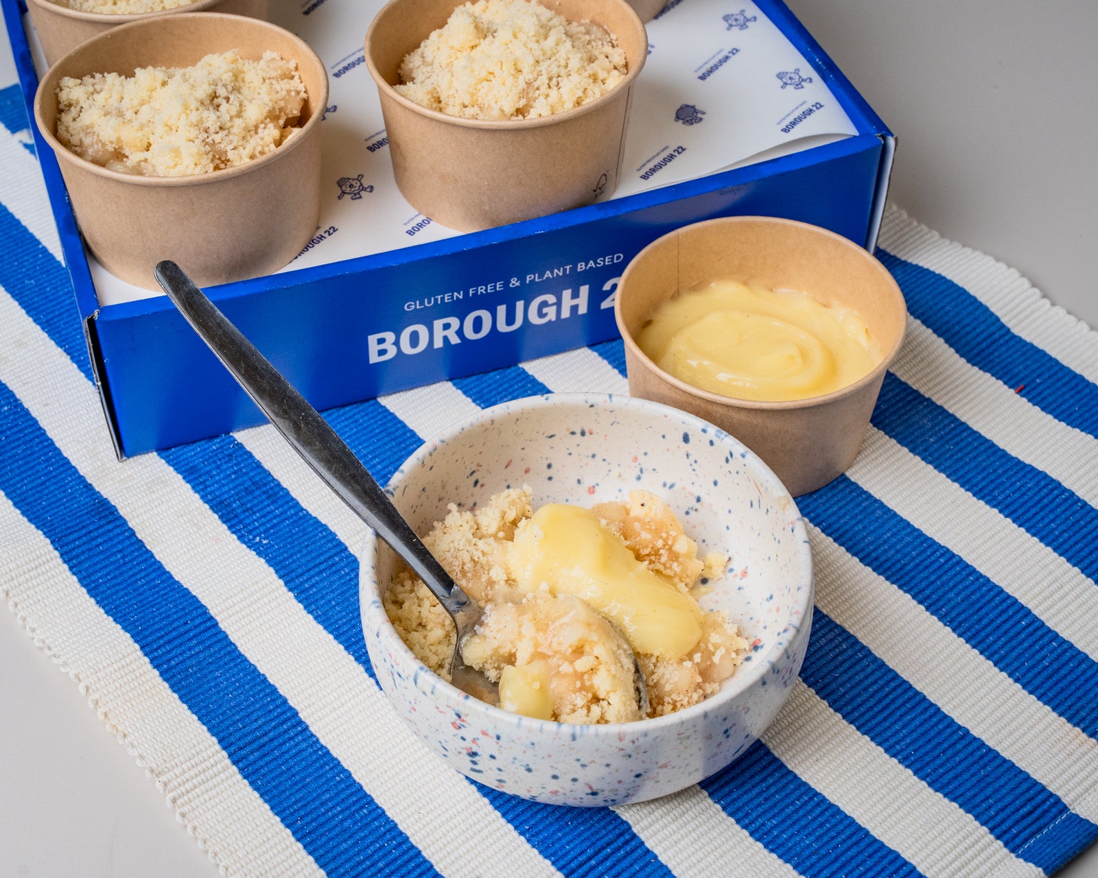 Borough 2 gluten free plant-based dessert cups shown with their blue branded box, arranged on a blue and white striped placemat. One cup is in the foreground topped with custard, and another plain cup sits beside it.