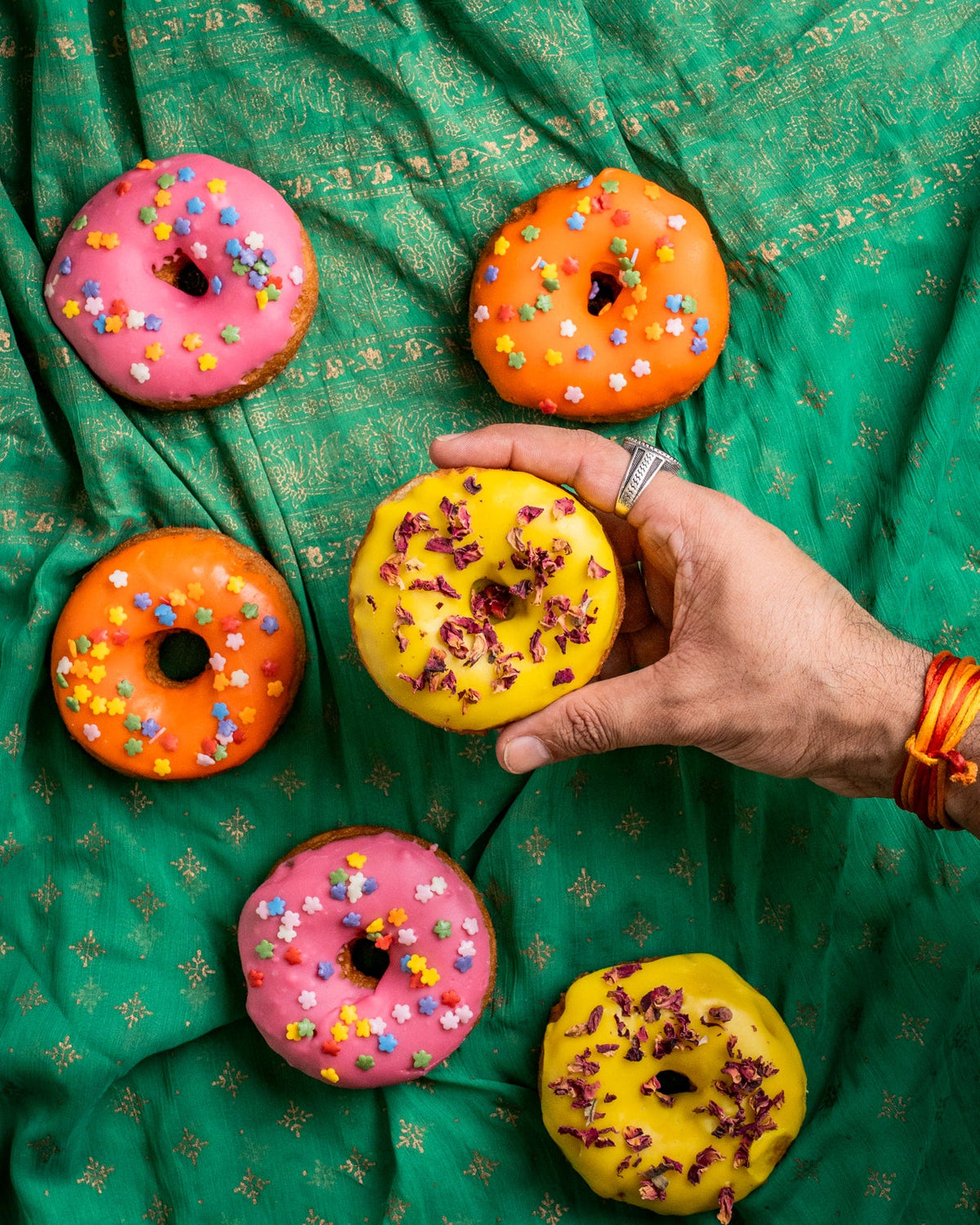Gluten free vegan doughnuts with bright pink, orange and yellow glazes, star sprinkles and rose petals on a green silk backdrop