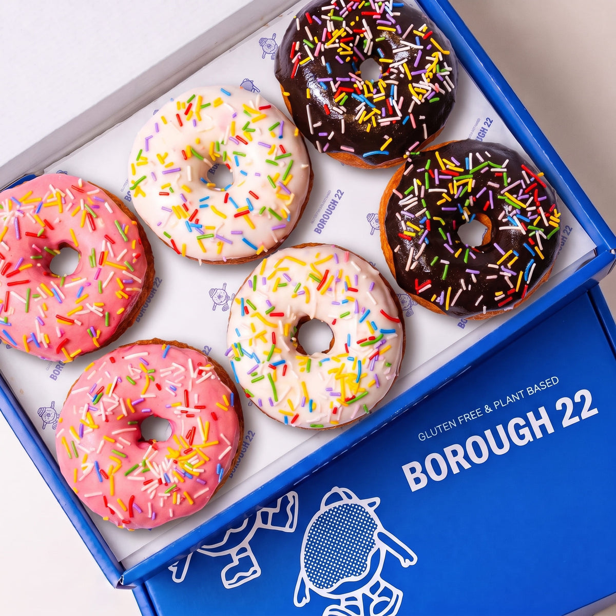 Open Borough 22 box of six gluten free oat free vegan fried ring doughnuts iced in pink, white and chocolate, all covered in rainbow sprinkles